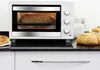 A white countertop oven with a loaf of bread baking inside, next to a loaf of bread and a jar of sugar. Produktbild-Vorschau