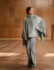 A woman models a gray knit poncho and matching pants in a room with wood-paneled walls and a wooden floor. Produktbild-Vorschau