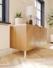 A wooden cabinet with a chevron pattern on its doors, standing on white legs, is placed in a room with a herringbone wooden floor. A white lamp with a wooden base and a potted plant are on top of the cabinet. Produktbild-Vorschau