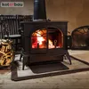 A wood-burning stove with a fire burning inside, placed on a wooden floor with a basket of firewood next to it. Produktbild-Vorschau