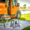 An orange camper van is parked next to a body of water, with outdoor furniture set up on a rug in front of it. Náhľad obrázku produktu