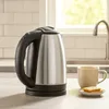 A stainless steel electric kettle is shown on a kitchen counter next to a mug of tea and cookies. Náhled obrázku produktu