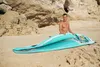 A man is sitting on a sandy beach behind an inflatable paddleboard. Produktbild-Vorschau