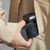 A person is holding a camera with a flash attached, tucking it into the pocket of a beige jacket. Podgląd zdjęcia produktu