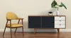 A yellow chair and a wooden sideboard with black and white drawers are placed against a cream-colored wall. Produktbild-Vorschau