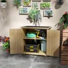 A wooden garden cabinet with its doors open, displaying gardening tools and supplies inside. The cabinet is placed on a tiled floor against a wall decorated with hanging succulent plants. Náhľad obrázku produktu