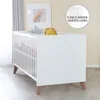 A white baby crib with wooden legs, decorated with stuffed toys and a framed picture on the wall. Produktbild-Vorschau