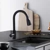 A modern black kitchen faucet is shown on a black countertop next to a sink. In the background, there are wooden cutting boards and a bowl of nuts. Produktbild-Vorschau