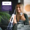 A person is holding a white water flosser with a clear reservoir filled with water, with other glasses and a plant in the background. Produktbild-Vorschau