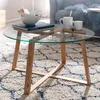 A round glass coffee table with wooden legs, decorated with books, a bowl, and mugs, placed on a patterned rug in a living room setting. Náhľad obrázku produktu