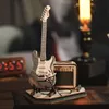 A miniature wooden model of an electric guitar and amplifier on a dark surface, with a blurred background featuring a lamp and furniture. Voir les images du produit