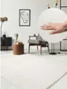 A hand touching a soft white rug in a modern living room setting. Produktbild-Vorschau