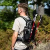 A person wearing a backpack with hiking poles attached, standing in a forest. Náhľad obrázku produktu