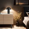 A bedside table with a lamp and flowers next to a bed. Voir les images du produit