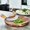 A kitchen counter scene with a mezzaluna knife and cutting board in the foreground, and a meal on a plate in the background. Anteprima dell’immagine prodotto