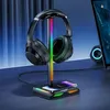 A black headphone stand with RGB lighting, holding a pair of black headphones, and a smartphone on a wireless charging pad, placed on a desk next to a laptop. Podgląd zdjęcia produktu