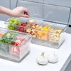 A hand is shown reaching into a clear plastic container filled with various fruits and vegetables, with two garlic bulbs in the foreground. Produktbild-Vorschau