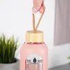 A hand holding a pink glass water bottle with a wooden lid and a woven strap. Voir les images du produit