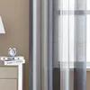 A close-up of a bedside table with books and an alarm clock, next to a window with sheer striped curtains. Produktbild-Vorschau