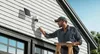 A man is installing a security camera on the side of a house. Náhľad obrázku produktu