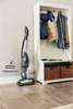 A vacuum cleaner is shown in a hallway next to a coat rack with jackets and leashes hanging on it. Voir les images du produit