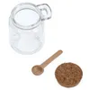 A clear glass jar with a handle, a wooden spoon, and a cork lid are arranged on a white background. Anteprima dell’immagine prodotto