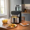 An air fryer is shown on a wooden table in a kitchen setting, with food items like french fries, skewers, and a salad nearby. Produktbild-Vorschau