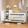 A white shoe cabinet with a padded seat and gold metal legs, featuring two flip-down shoe storage compartments and a side cabinet. It is placed on a wooden floor in a hallway setting, with a yellow handbag and coats hanging on the wall. Produktbild-Vorschau