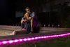 A couple is sitting on wooden steps outdoors at night, illuminated by pink LED lights. Produktbild-Vorschau