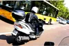A person riding a white scooter in traffic with a yellow bus in the background. Anteprima dell’immagine prodotto