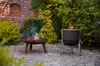 Two fire pits are shown in a garden setting with a brick wall and lush greenery. Produktbild-Vorschau