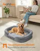 A golden retriever dog lies on a gray dog bed in a living room, with a woman sitting on a sofa in the background reading a book. Náhled obrázku produktu