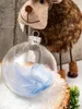 A clear glass Christmas ornament filled with blue and white feathers sits on a snowy surface next to a wooden log. A fuzzy reindeer decoration is in the background. Podgląd zdjęcia produktu