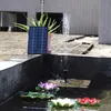 A solar-powered fountain pump is shown in a small pond with artificial lotus flowers, with a solar panel and a potted plant in the background. Náhled obrázku produktu