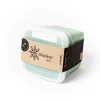 A light green lunchbox with a white lid, wrapped in a paper band with branding and product information. Produktbild-Vorschau