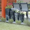 A set of tall, dark planters with various flowers and greenery arranged on a patio next to outdoor chairs. Produktbild-Vorschau