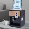 A mini desktop water dispenser with a water bottle on top, placed on a table next to a cup and spoon. A sofa is visible in the background. Náhled obrázku produktu