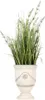 A potted artificial plant with green grass-like leaves and small purple flowers in a decorative beige urn-shaped pot. Podgląd zdjęcia produktu