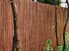 A close-up view of a woven bamboo fence with trees and plants in the foreground and background. Produktbild-Vorschau