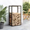 A black metal firewood rack filled with logs is placed on a patio next to a white wall and a potted plant. A wooden chair and table are visible in the background. Náhľad obrázku produktu