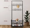 A black metal coat rack with shelves for shoes, a hat, and a bouquet of flowers on the middle shelf, placed against a light gray wall with a potted plant to the right. Produktbild-Vorschau