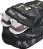 A close-up view of a black camouflage backpack with gold Under Armour logos, showing a pair of athletic shoes peeking out of a zippered compartment. Náhled obrázku produktu