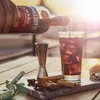A hand pours Ramazzotti Amaro from a bottle into a glass filled with ice and a dark liquid, with spices and a jigger on a marble board. Produktbild-Vorschau