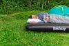 A man sleeping on an air mattress next to a tent in a grassy, wooded area. Náhled obrázku produktu