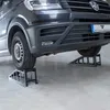 A dark gray van is parked on a concrete floor, with its front wheels resting on two black ramps. The ramps are angled and have a slatted design. A license plate with a logo is visible on the front of the van. Podgląd zdjęcia produktu