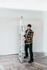 A man is holding a folded ladder in a room that appears to be under renovation, with plastic sheeting on the floor. Náhled obrázku produktu