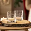 Two clear glasses with festive etchings sit on a rustic wooden slice next to a lit candle. Produktbild-Vorschau