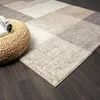 A close-up view of a rug with a geometric pattern, placed on a dark wooden floor next to a woven ottoman with a magazine on it. Produktbild-Vorschau