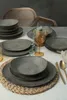 A table setting with stoneware plates, bowls, and a glass of amber liquid, with chairs in the background. Anteprima dell’immagine prodotto