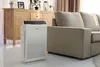 An air purifier is placed on a white floor in a living room, next to a sofa and a wooden cabinet. Produktbild-Vorschau